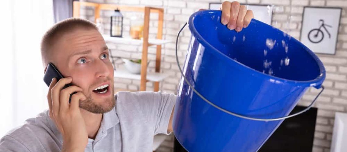 Young Man Collecting Water Leakage In Bucket While Calling Plumber On Smartphone